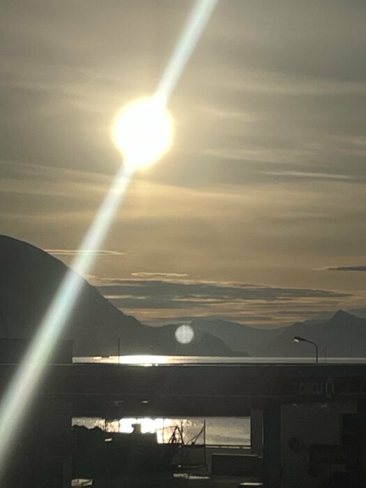 Solnedgång över ett bergslandskap med reflektioner i vattnet och silhuetter av byggnader.