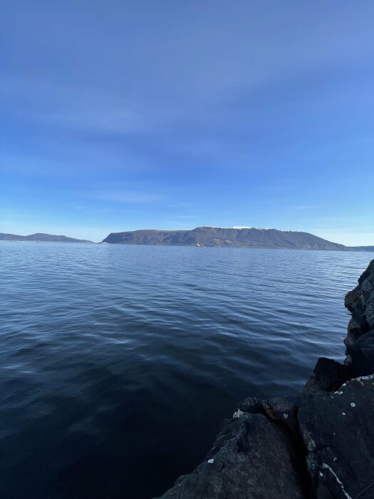 Utsikt över en lugn sjö med klippor i förgrunden och berg i bakgrunden under en klarblå himmel.