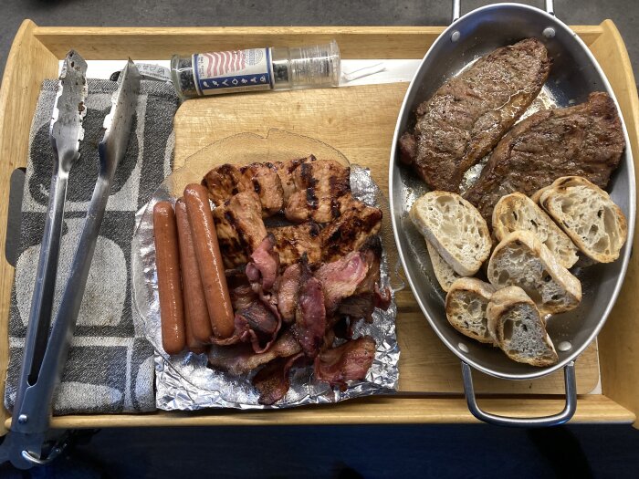 Grillat kött, korv och bacon på ett serveringsfat bredvid rostat bröd och grilltillbehör.