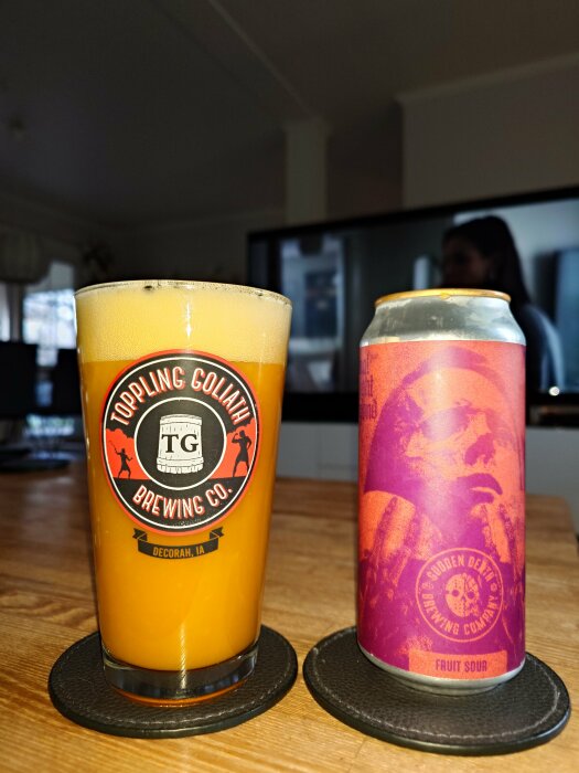 Ölglas med orange färgat skum bredvid en ölburk från Toppling Goliath Brewing Co., bakgrunden visar en suddig inomhusmiljö.