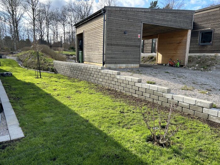 Befintlig mur vid hus och gräsmatta som ska bytas ut, synligt grävd mark och barnets cykel vid carport.