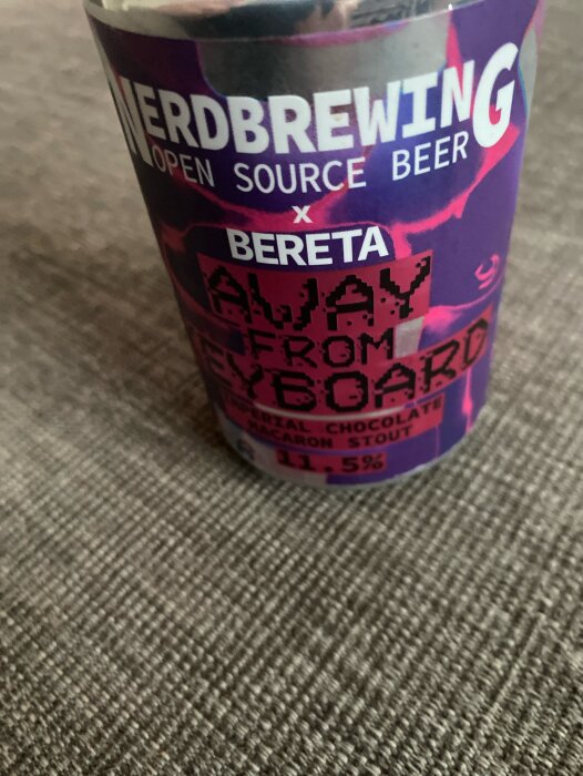 En ölburk med texten "Nerdbrewing x Bereta Away From Keyboard Imperial Chocolate Macaron Stout" på en textilbakgrund.