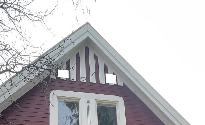 Vitdekorerade gavelspetsar på rödmålat hus med vit fönsterram och grenar i förgrunden.