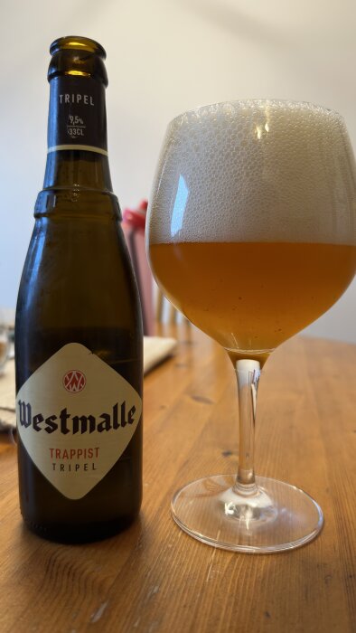 Flaska Westmalle Trappist Tripel bredvid ett glas med skummande öl på ett träbord.