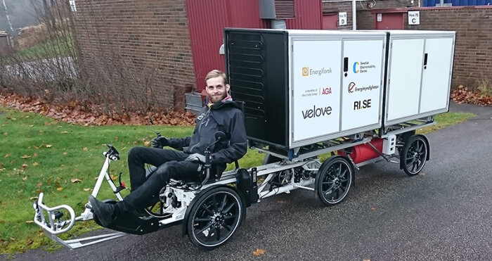 Person sitter på en fyra-hjulig transportcykel med stora förvaringslådor på ett industriområde.