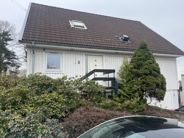 Husfasad med sliten vit ytterdörr och svart trappräcke, omgiven av grönska och en framskjuten garagetakskant.
