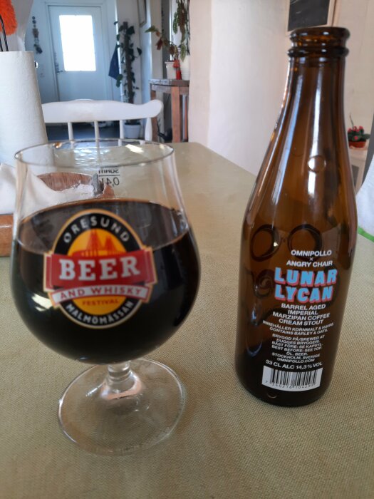 Ett glas mörk öl och en flaska av Omnipollo Lunar Lycan på ett köksbord.