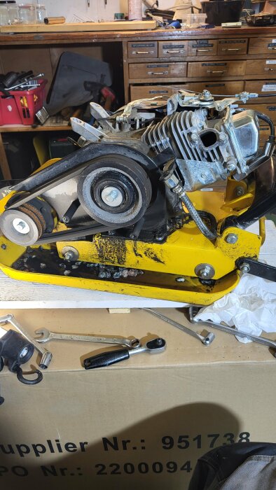 Öppen motor på en gul markvibrator med synlig oljeläckage och verktyg runtomkring i en verkstadsinställning.