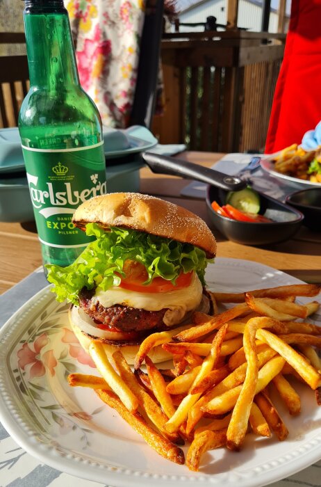 Högrevsburgare med sallad och tomat på tallrik bredvid pommes frites och en Carlsberg ölflaska, utomhus på altan.