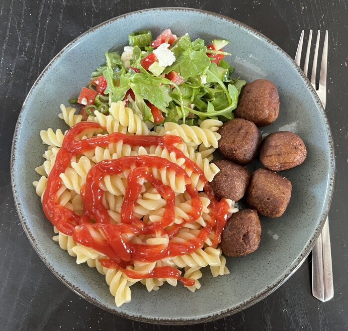 Tallrik med kycklingköttbullar, skruvpasta, sallad och ketchup.