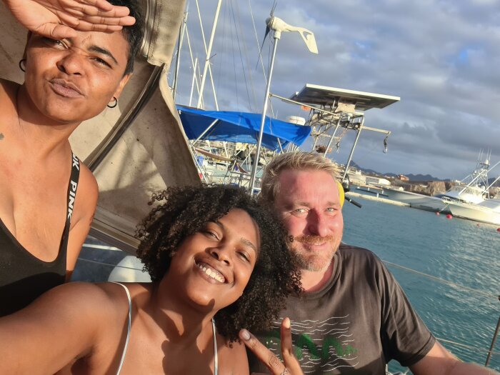 Tre personer som tar en selfie på en båt med segelbåtar och hav i bakgrunden.