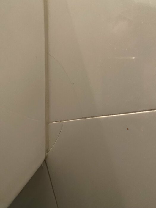 Två sprickor i vita kakelplattor på badrumsvägg nedanför en vägghängd toalett.
