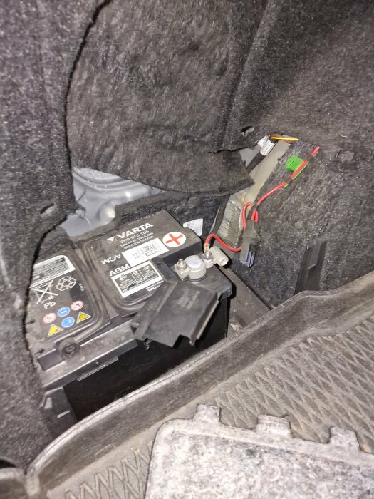 Bilbatteri placerat till vänster i ett bilens bagageutrymme med synliga anslutningar och kablar.