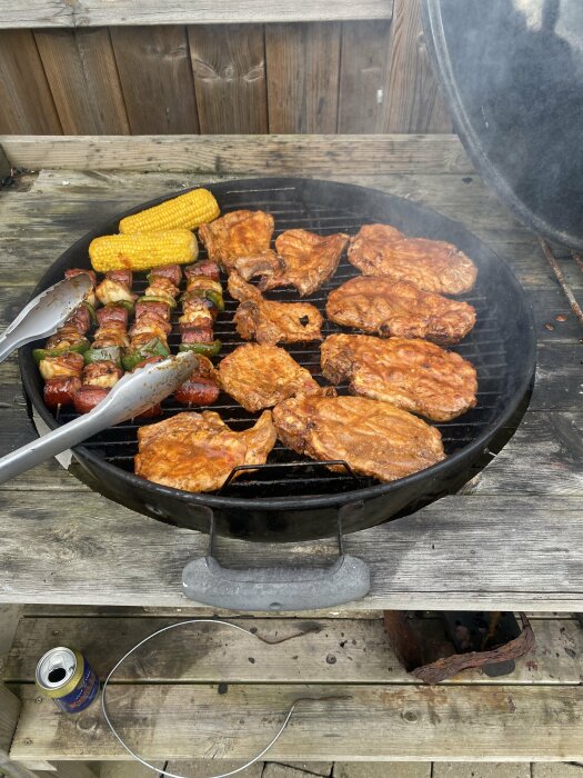 Rostig Weber-grill med kött, majs på kolven och grönsaks-korvspett på ett trädeck.