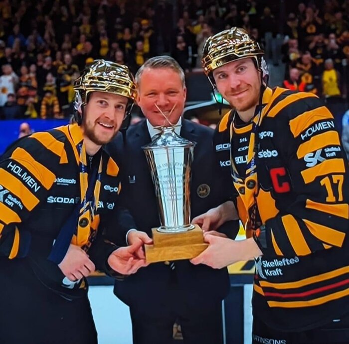 Tre personer i ishockeydräkter håller i en pokal, glad stämning och segerfirande.