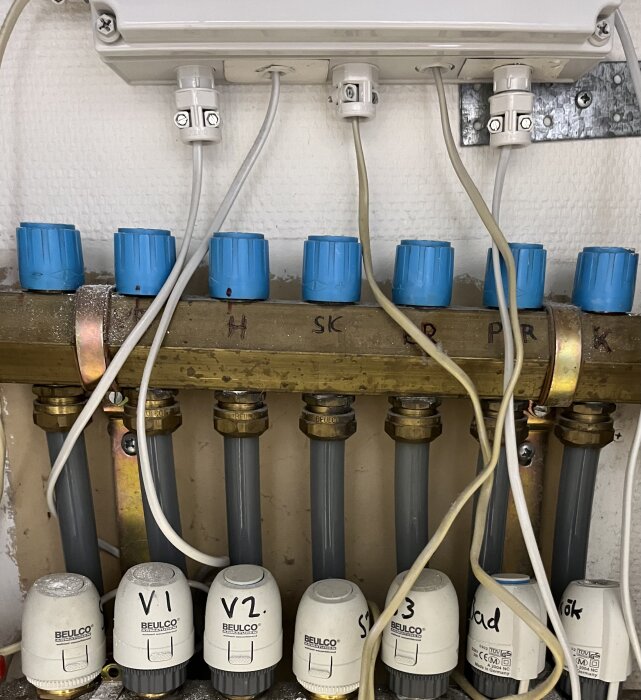 VVS-installation med rördelare, termostater och kopplingar på en vit vägg.