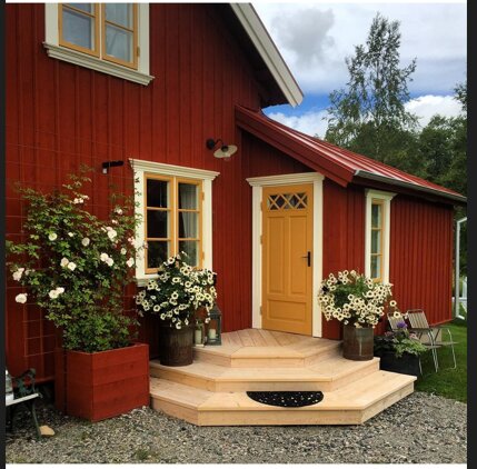 Halvmåneformad entrétrappa i trä framför rött hus med gult dörr och blommande buskar.