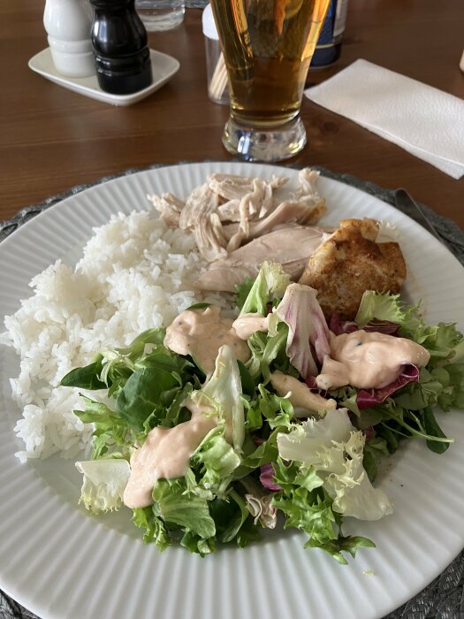 Tallrik med ugnsstekt kyckling, ris, sallad och dressing, samt ett glas öl i bakgrunden.