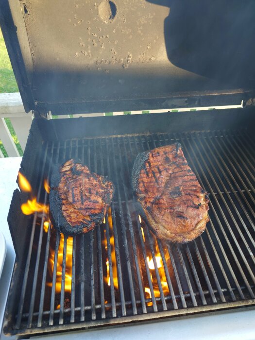 Två biffar grillas på en smutsig gasolgrill med synliga lågor under gallret.