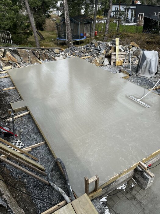 Ny gjuten betonggrund för pool med pumpslang och formar omgiven av grus och stenar.