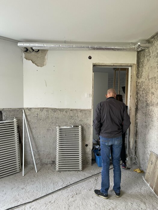 Renoveringsarbete i rum med synligt isolerat avloppsrör och radiatorelement, person står och tittar.