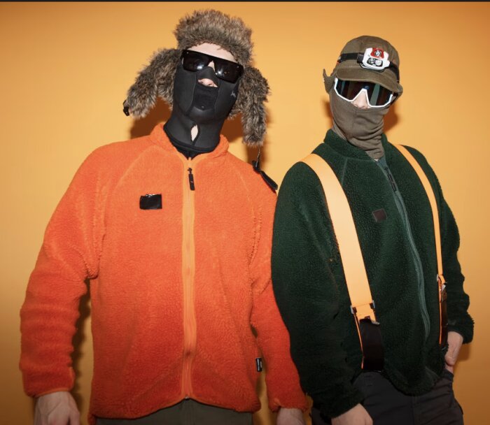 Två personer i varma jackor och ansiktsmasker står framför en orange bakgrund.