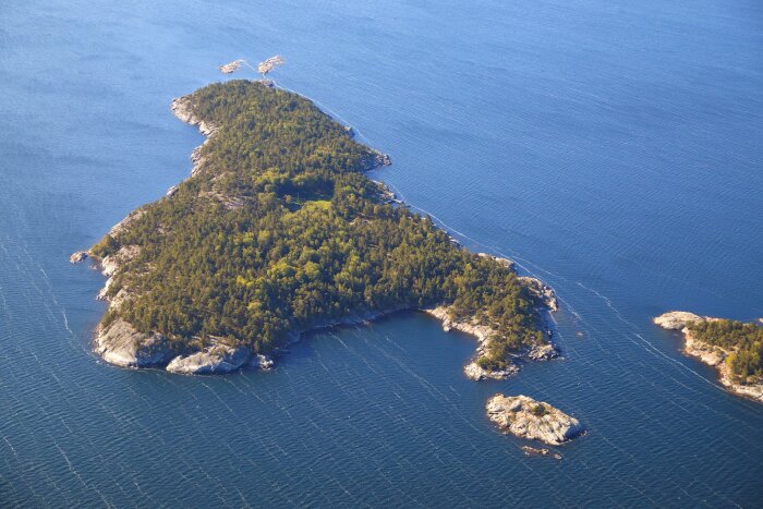 Flygbild över en ö med tät skog och klippiga stränder i skärgården vid Smådalarö.