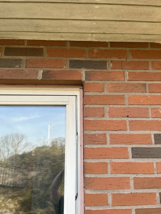Tegelfasads hörna med sprickor nära taket ovanför ett fönster, ingen synlig skada på lägre tegel eller fog.