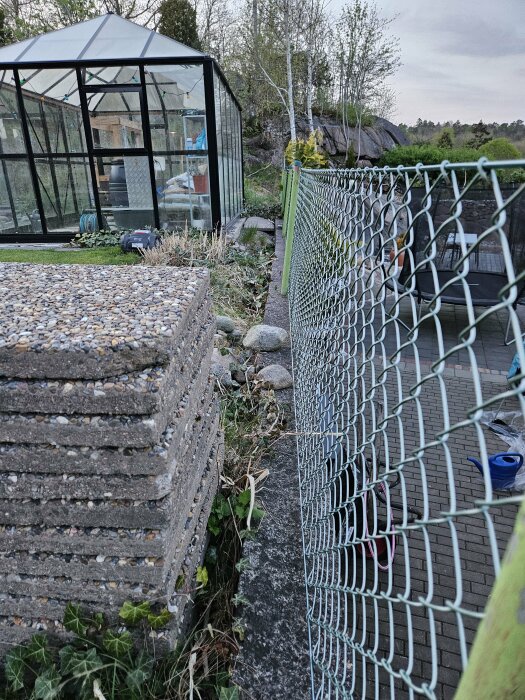 Gammal betongstödmur som lutar utåt intill ett staket och växthus med synligt markförskjutning.