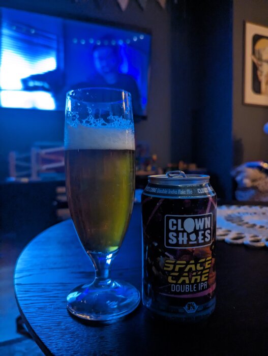 Ett glas öl bredvid en burk med texten 'CLOWN SHOES SPACE CAKE DOUBLE IPA', i ett mörkt rum med en TV i bakgrunden.