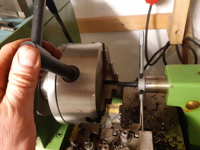Hand som håller i en axel i en svarv med gängad metallstång och svarvverktyg synliga, indikerar verktygstillverkning.