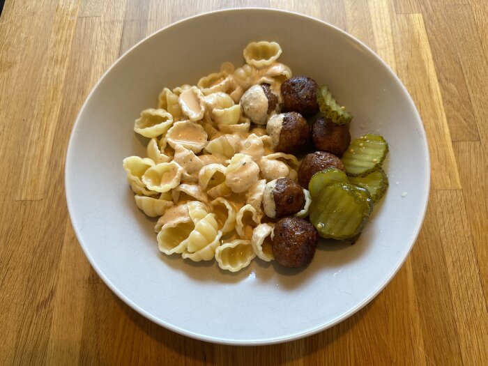 Skål med pasta, köttbullar och skivade inlagda gurkor på träbord.