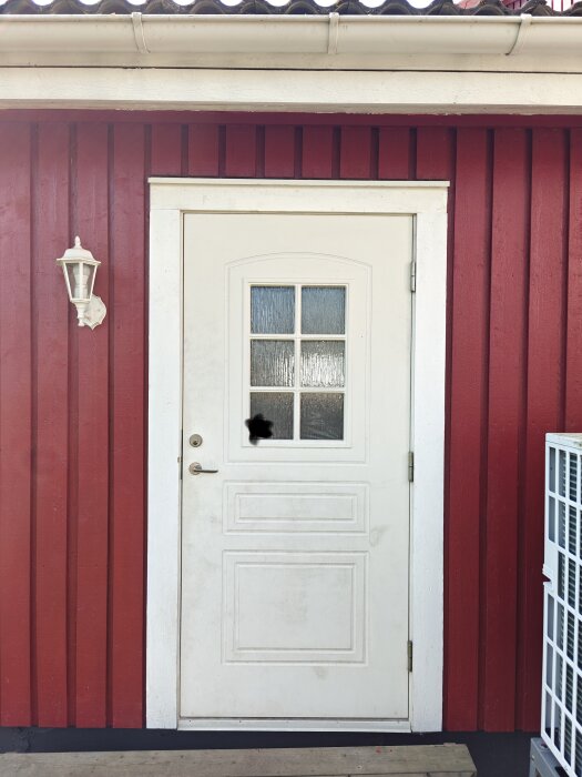 Vit entrédörr i en röd träfasad utan taköverbyggnad och med ett ljus bredvid dörren.