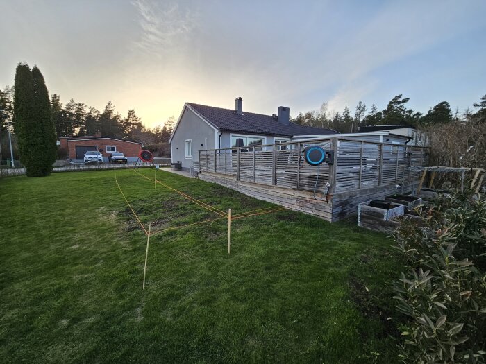 Trädgård med befintlig trädäck och planerad utbyggnad markerad med snören, hus och luftvärmepump i bakgrunden.