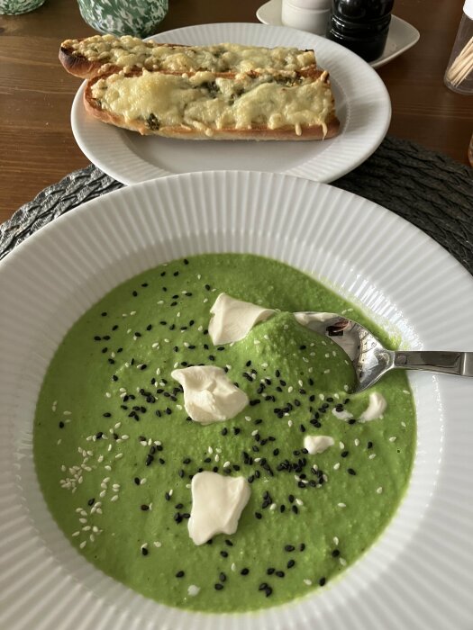 En skål med ärtsoppa toppad med gräddklickar och svarta sesamfrön, bakom en ostgratinerad baguette.