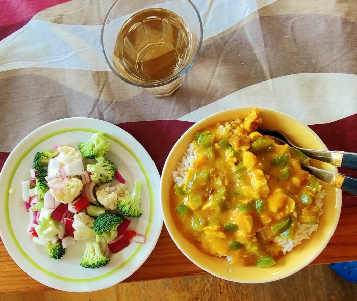 Chicken à la king över ris bredvid tallrik med snabbpicklad sallad, glas vatten ovanpå randig duk.