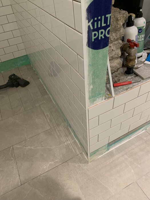 Hörn av ett badrum under renovering med vita kakelplattor och byggmaterial på golvet.