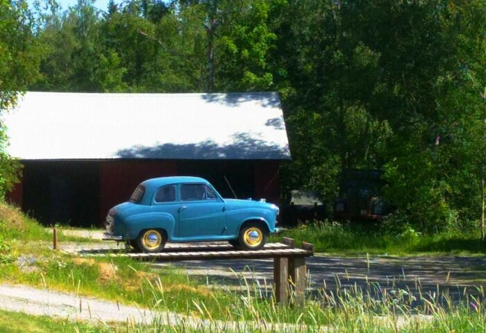En blå Austin A35 parkerad på en smörjbrygga framför ett lantligt garage omgivet av grönska.