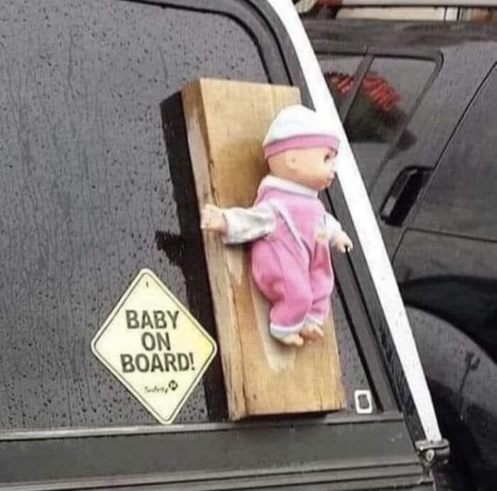 Docka iklädd rosa kläder fäst vid bilfönster bredvid en "Baby on board"-skylt.