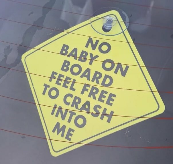 Gul skylt med texten "NO BABY ON BOARD FEEL FREE TO CRASH INTO ME" på en bilruta.