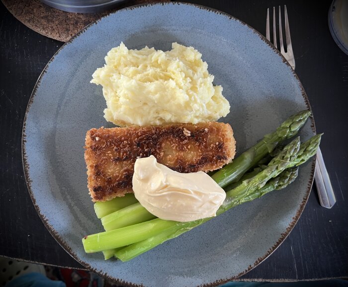 Panerad torsk, mosad potatis, kokt sparris och klick hollandaisesås på mörkgrå tallrik.