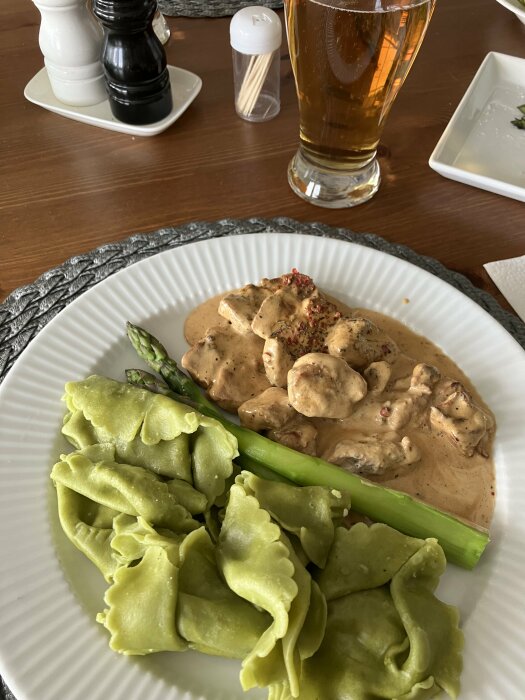 Fläskfilé i gräddsås med kryddor, gröna spenat- och ricottafyllda pastakuddar, kokt sparris och ölglas i bakgrunden.