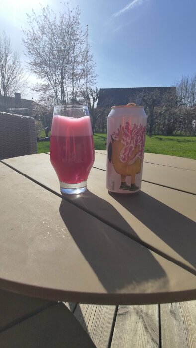 Ett glas med rosa dryck och en burk med färgglad etikett på ett utebord i solen.