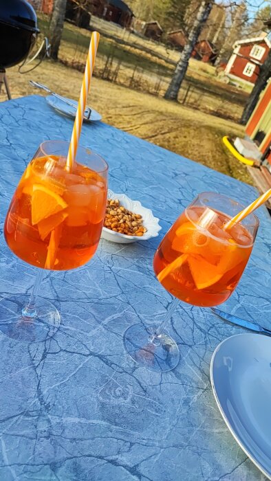Två glas med Aperol Spritz och en skål med nötter på ett blått bord utomhus.