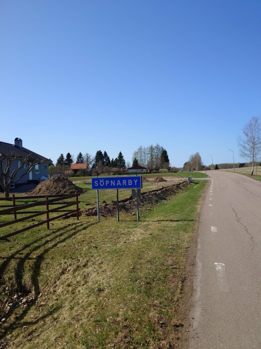 Landsväg med ortnamnsskylt "SÖPNARBY", grönt gräs och blå himmel, några träd och en villa.