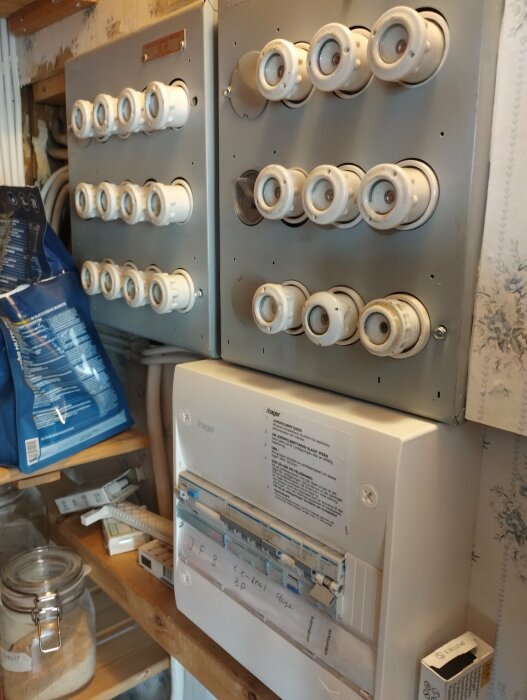Elektriskt säkringsskåp med flera säkringsomkopplare och modern elcentral i ett utrymme med tapeter och hyllor.