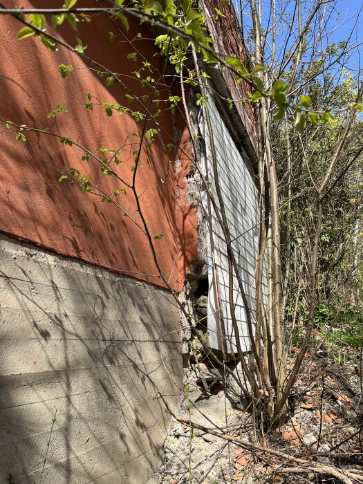 Exponerad isolering på halvfärdig fasad av tvåvåningshus, med synliga skador och vegetation.