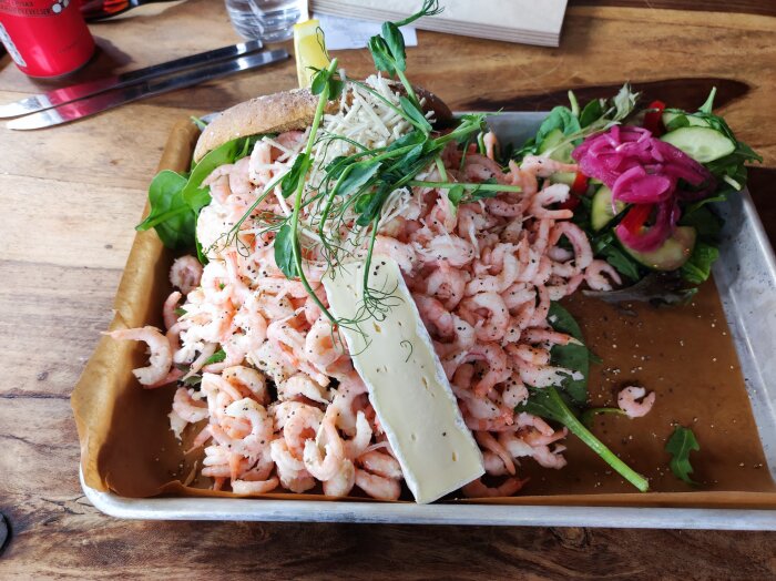 Räkmacka med generöst med handskalade räkor, sallad, dill, citron och en skiva brieost på ett träbord.