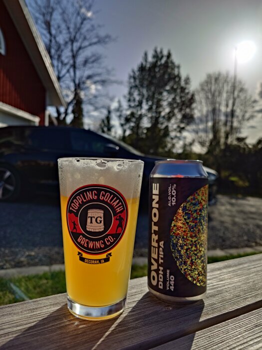 Ölglas från Toppling Goliath Brewing Co. och en Overtone-öldosa på ett träbord utomhus i solljus.