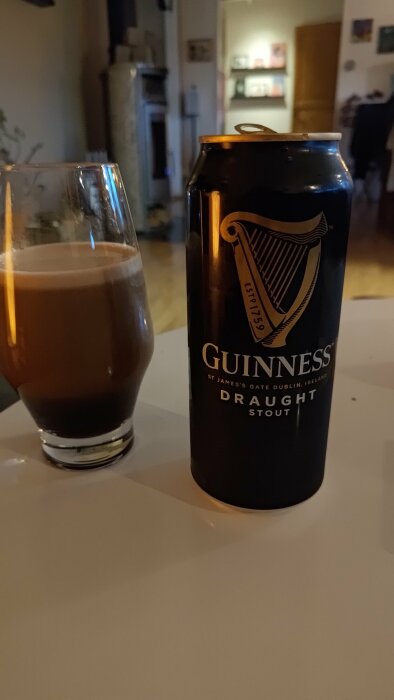 En burk Guinness öl bredvid ett halvfullt glas på ett bord i ett suddigt upplyst vardagsrum.